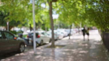 Park halindeki arabaların ve yeşil ağaçların arasında güneşli bir şehir ortamında yürüyen insanların olduğu bir açık şehir sokağının bulanık arka planı.