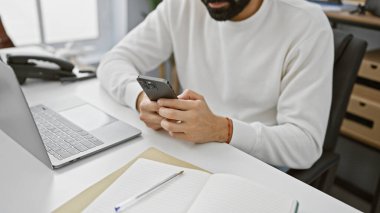 Modern ofis masasında dizüstü bilgisayarı ve defteri olan akıllı telefon kullanan İspanyol profesyonel bir adam..