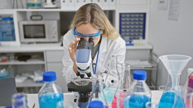 Modern bir laboratuarda mikroskop kullanan laboratuvar önlüklü ve koruyucu gözlüklü genç bir kadın..