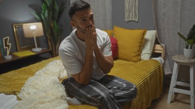 Beyaz gömlekli ve ekoseli sakallı yakışıklı İspanyol bir adam modern dekorlu rahat bir yatak odasında hardal yatağında oturuyor ve gece evde dalgın dalgın görünüyor..