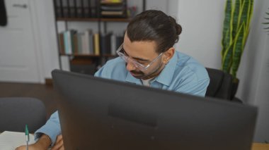 Ofiste çalışan genç İspanyol bir adam, bir not defterine notlar yazarken bilgisayar ekranına odaklanıyor.