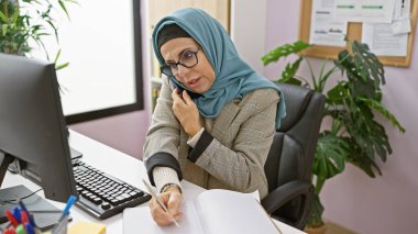 Modern bir ofiste tesettürlü profesyonel bir kadın telefonda notlar yazarken birden fazla iş yapıyor..