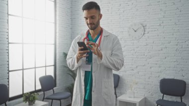 Genç İspanyol erkek doktor, hastane bekleme odasında akıllı telefon kullanıyor. Boynunda steteskop ve kendine güvenen bir gülümseme modern bir sağlık hizmetinin göstergesi..