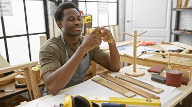 Afro-Amerikalı bir adam, marangozluk atölyesinde kahve molası veriyor. Etrafı ahşap işçiliği aletleri ve projelerle çevrili..