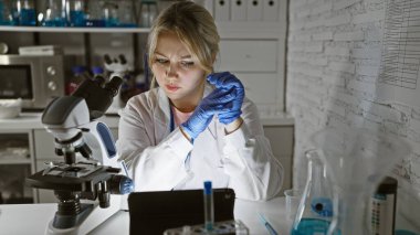 Genç bir kadın bilim adamı, kapalı bir laboratuvar ortamında, ekipmanlarla çevrili bir tabletin verilerini analiz ediyor..