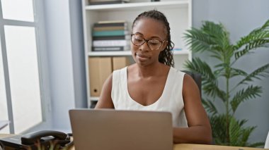 Ofis masasında çalışan gözlüklü, modern dekorla çevrili Afro-Amerikan kadın..