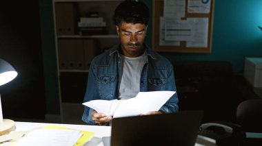 Odaklanmış, sakallı İspanyol bir adam geceleri iyi aydınlatılmış bir ofis ortamında belgeleri inceler, iş ve konsantrasyon tasvir eder..