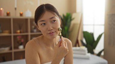 Young chinese woman in a spa room using a facial roller for skincare enhancement in a serene wellness setting clipart