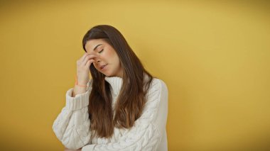 Beyaz kazaklı stresli genç bir İspanyol kadın sarı arka planda baş ağrısı ile poz veriyor..