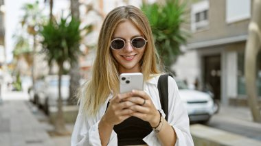 Güneş gözlüklü şık bir genç kadın güneşli bir şehirde akıllı telefon kullanıyor..