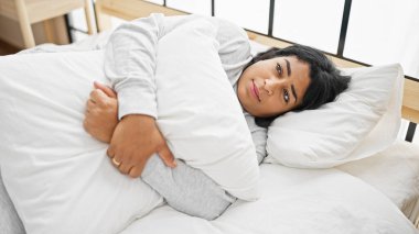 Düşünceli bir İspanyol kadın, parlak bir yatak odası ortamında yastığı kucaklıyor. Düşünceyi ya da üzüntüyü sembolize ediyor..