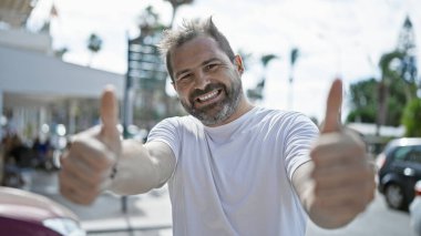 Yakışıklı, olgun, gri saçlı bir İspanyol açık havada güneşli bir caddede başparmağını sallıyor..