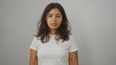 Çekici genç İspanyol kadın izole edilmiş beyaz arka planda dikiliyor günlük beyaz bir tişört giyiyor ve ciddi görünüyor..