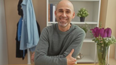 Yakışıklı, kel, sakallı, rahat bir evde gülümseyen, süveter giyen bir adam..