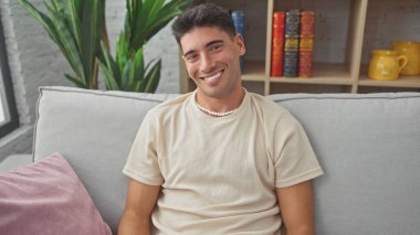 Yakışıklı İspanyol adam rahat bir oturma odasında kanepede otururken gelişigüzel gülümsüyor..