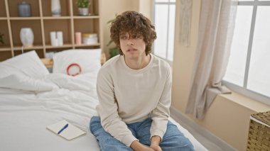 Dalgın, kıvırcık saçlı, yatakta oturan genç bir adam. Yanında bir defter, modern bir yatak odasında.