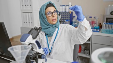 Orta yaşlı tesettürlü bir kadın modern bir laboratuvar ortamında deney tüpünü inceliyor. Bilim alanında çeşitlilik gösteriyor..