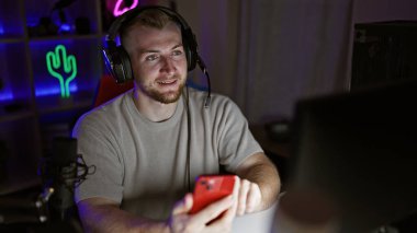 Akşamları karanlık bir oyun odasında akıllı telefon ve kulaklık kullanan sakallı yakışıklı beyaz adam.