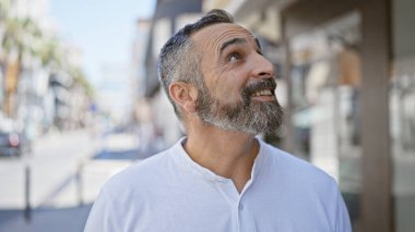 Sakallı, gri saçlı neşeli, olgun bir İspanyol, şehir caddesinde dikilirken kafasını kaldırıyor..