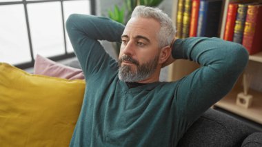 Rahat sakallı adam renkli yastıklar ve kitaplarla dolu bir oturma odasında dinleniyor..