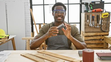 Güvenlik gözlüğü takan gülümseyen Afrikalı adam güneşli bir atölyede tahtayı inceliyor..