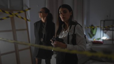 Two women investigators with badges and camera inspect a dimly lit indoor crime scene surrounded by caution tape. clipart