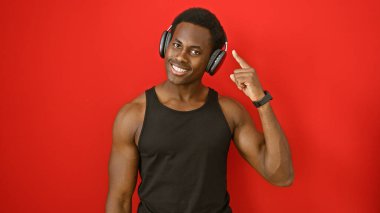 Siyah kolsuz tişörtlü gülümseyen Afro-Amerikan adam kulaklık takıyor ve kafasını kırmızı arka plana doğrultuyor..