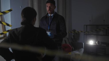 Dedektifler, bir kadın ve bir erkek, kapalı bir suç mahallindeki ipuçlarını dikkatli bir bant, kanıt işaretleyicileri, dramatik ışıklandırma ve karanlık ortam ile analiz ederler..