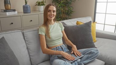 Genç sarışın bir kadın modern bir oturma odasında oturuyor. Çiçekler ve yastıklarla, kameraya gülümsüyor..