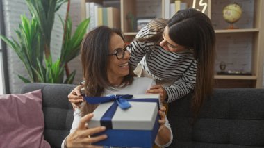 Bir anne ve kızı, sıcak bir oturma odasında bir hediyeyle neşeli bir anı paylaşıyor. Aile sevgisini ve birlikteliği temsil ediyor..