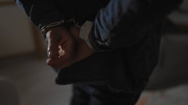 A handcuffed man's hands close-up in a dimly lit indoor scene portraying arrest or detainment. clipart