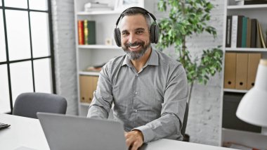 Ofiste dizüstü bilgisayar kullanarak kulaklık takan gülümseyen, kıdemli İspanyol sakallı adam..