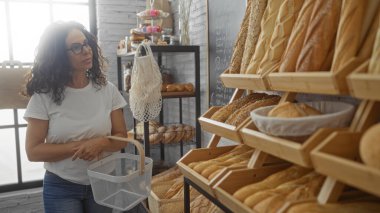 Ahşap raflarda sergilenen çeşitli ekmek çeşitleriyle dolu bir fırında alışveriş yapan bir kadın.