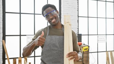 Güvenlik gözlüklü gülümseyen marangoz iyi aydınlatılmış bir atölyede başparmak kaldırıyor.