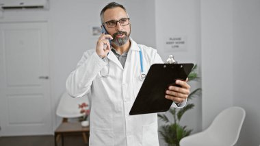 Latin asıllı kıdemli erkek doktor elinde dosyayla telefonda konuşuyor..