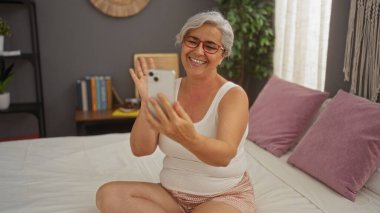 Gri saçlı olgun bir kadın yatak odasında video çağrısı yapıyor. Yatağında oturmuş gülümsüyor ve el sallıyor..