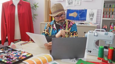 Gözlüklü ve sakallı şık bir adam dizüstü bilgisayarda kumaş ve dikiş malzemeleriyle çevrili renkli bir terzide çalışıyor..