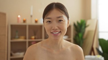 Yumuşak ışıklandırma ve doğal dekor unsurlarıyla huzurlu bir spa odasında gülümseyen genç Çinli kadın, sakinlik ve sağIık hissi uyandırıyor..