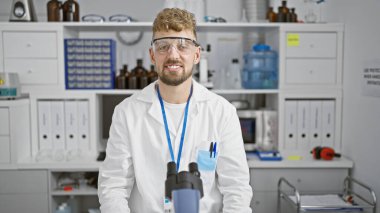 Mavi gözlü, sakallı, güvenlik gözlüğü takan ve laboratuvar önlüklü beyaz bir adam iyi donanımlı bir laboratuarda duruyor..