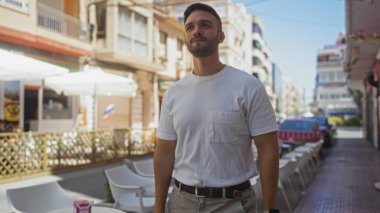 Şehirdeki bir sokakta beyaz gömlek giymiş, sakallı yakışıklı İspanyol bir adam. Arka planda açık kafe ve binalarla çevrili bir kasaba..