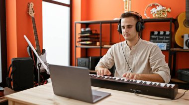 Odaklanmış bir adam renkli bir müzik stüdyosunda gitar ve kulaklıkla klavye çalar..