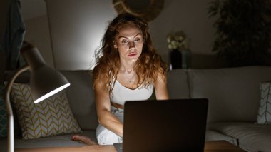 Geceleri rahat bir oturma odasında dizüstü bilgisayar kullanan odaklanmış genç bir kadın bir lamba ve çevre ışıkları ile aydınlatılıyor..