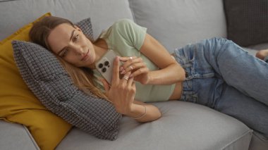 Sarışın, güzel bir kadın oturma odasındaki kanepede uzanmış, akıllı telefon kullanıyor, rahat ve rahat bir atmosfer sergiliyor..