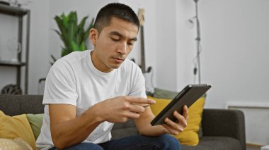 İspanyol adam modern oturma odasında tablet kullanıyor. Teknoloji ve yaşam tarzı sergiliyor..