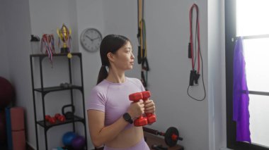 Kadın spor salonunda spor yapıyor. Arka planda kırmızı halterler ve spor aletleri var..