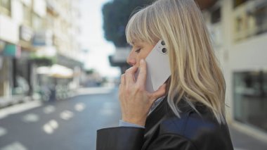 Sarışın bir kadın şehrin hareketli bir caddesinde dikilirken akıllı bir telefonla konuşuyor..