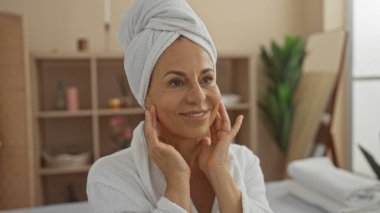 Olgun İspanyol kadın spa sağlık merkezinde rahatlatıcı bir anın tadını çıkarıyor, yüzünü nazikçe tutarken gülümsüyor, bornoz giyiyor ve havluyu içeride tutuyor.