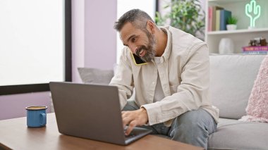 Yakışıklı, yaşlı, sakallı bir adam. Evdeki koltukta oturmuş telefonla konuşuyor..