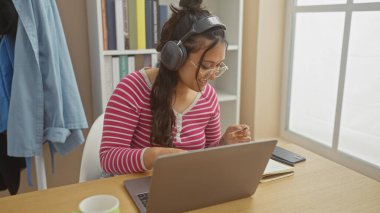 Evde dizüstü bilgisayarla çalışırken gülümseyen, kulaklık takan ve günlük giysiler giyen İspanyol bir kadın..