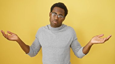 Kafası karışmış, gri kazaklı, sarı bir duvara karşı belirsizlik gösteren Afro-Amerikan bir adam..